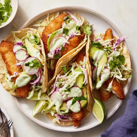 Close-up of Baja Fish Tacos topped with avocado, red onion, and zesty drizzle, served with lime wedges on a rustic table.