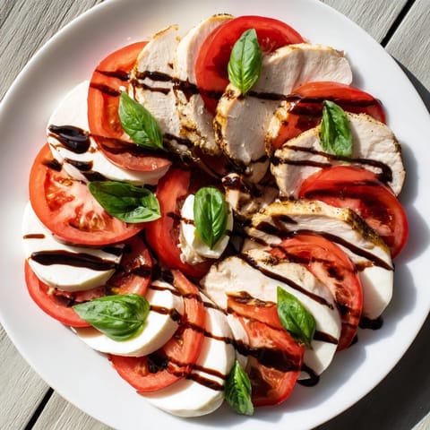 A close-up of Chicken Caprese Salad with layered ingredients, showcasing fresh basil leaves and a glossy balsamic drizzle on the hearty grilled chicken slices.